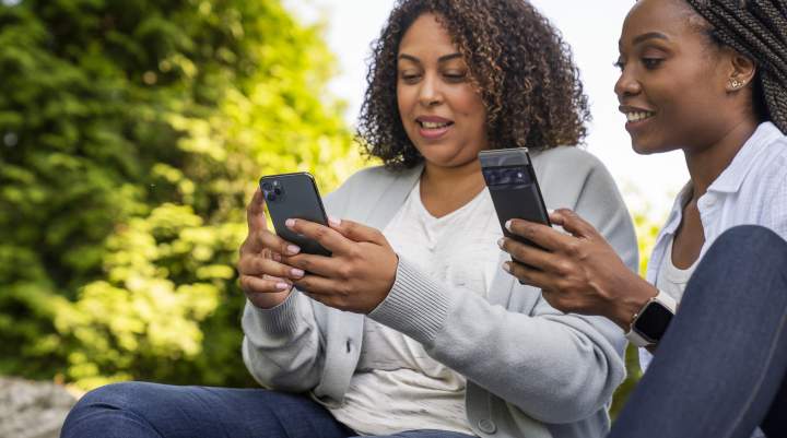 Two women sit outside together playing on their smartphones. 