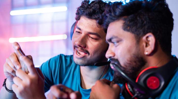 Two men wearing gaming headsets watch something on a smartphone.